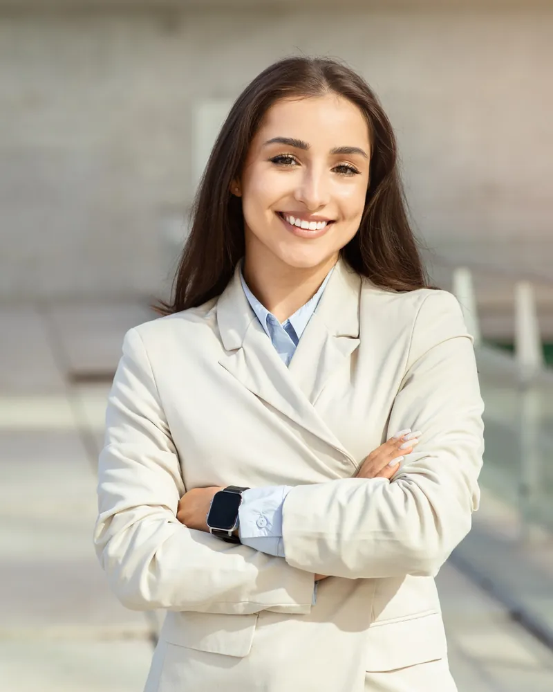 A smiling professional in a beige suit, arms crossed, outside an office building in soft daylight
