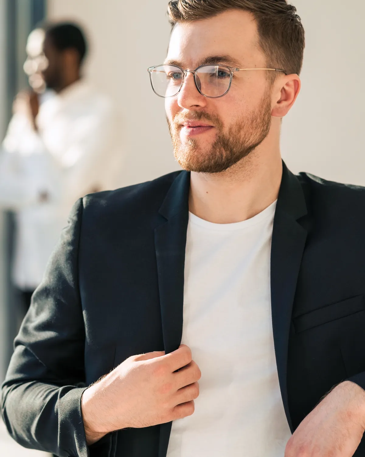 A professional in glasses and a navy blazer by a bright window, looking away from the camera, with colleagues talking out of focus behind him